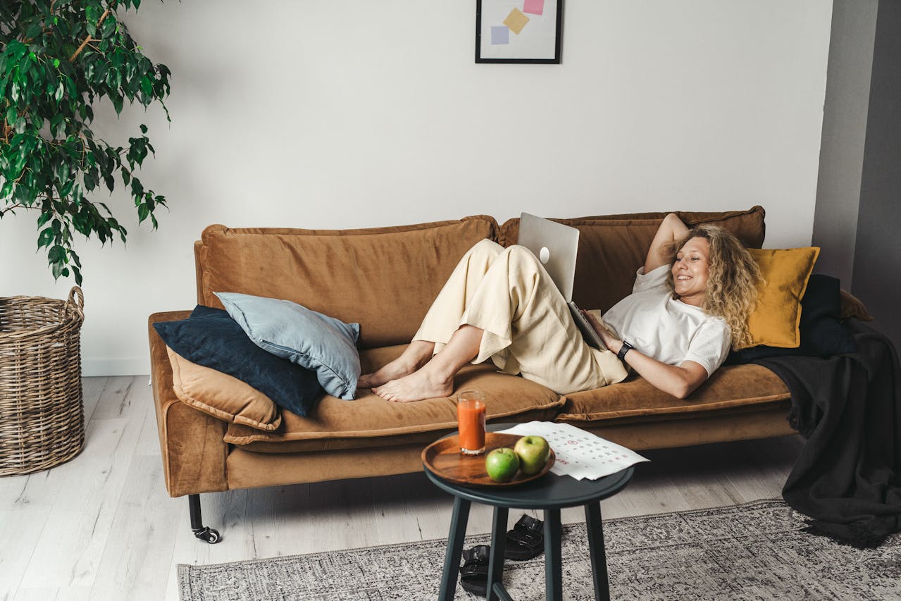 Vrouw ligt ontspannen met laptop op de bank, met een glas sap en fruit op tafel – een beeld van balans tussen werk en ontspanning.